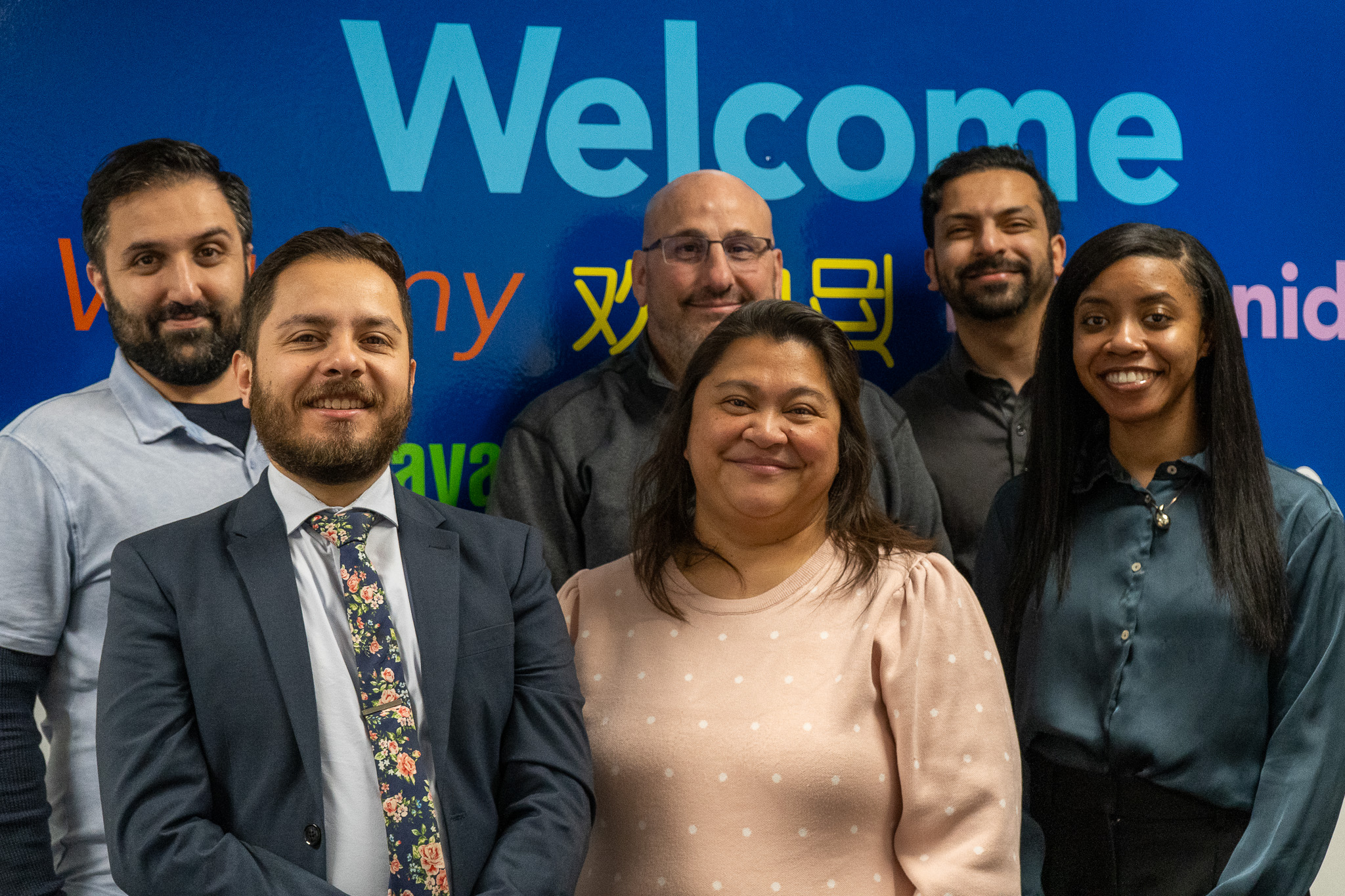 Six smiling diverse people standing in front of a blue "Welcome" sign.